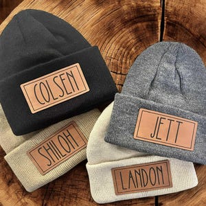 May include: Four knit beanies in black, gray, and beige, each with a rectangular leather patch. The patches display names: "COLSEN", "SHILOH", "JETT", and "LANDON" in a handwritten style font. The beanies are arranged on a wooden surface.