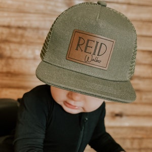 May include: An olive green trucker hat with a brown leather patch that reads "REID Walker" in white lettering. The hat is worn by a person wearing a black jumpsuit.