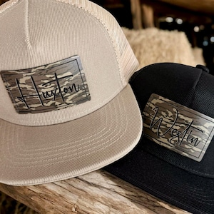May include: Two trucker hats, one tan and one black, are displayed. The tan hat has a beige mesh back and a rectangular patch with the name "Huxton" in a camouflage pattern. The black hat has a similar patch with the name "Westin".