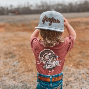 May include: A young child wearing a gray and brown baseball cap with the name "Grayson" embroidered on it. The child is also wearing a red t-shirt with a turkey graphic and the words "Southern Boy" printed on it. The child is wearing blue jeans with a brown belt.