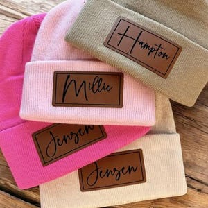May include: A stack of four beanies in shades of pink, tan, and cream. Each beanie has a brown leather patch with a name in a cursive font. The names are "Hampton", "Millie", and "Jensen". The beanies are folded over.