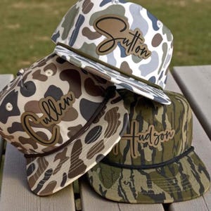May include: Three camouflage print baseball caps with names: "Sutton", "Cullen", and "Hudson". The caps have a flat brim and a brown leather patch with the name in a script font. The caps are stacked on a wooden surface.