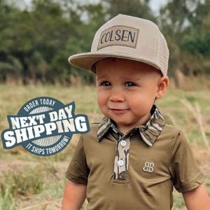 May include: A young child wearing a khaki baseball cap with the name "COLSEN" on the front. The child is also wearing a khaki polo shirt with a camouflage collar and button placket. The shirt has a small logo on the chest.