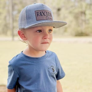 May include: A young child wearing a blue t-shirt with a small logo and distressed denim shorts. The child is wearing a gray and brown baseball cap with the text "RAXTON" on the front.