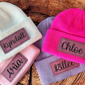 May include: A collection of personalized beanies in various colors, including white, pink, purple, and hot pink. Each beanie features a rectangular leather patch with a name printed on it. The names include "Kyndall", "Aria", "Chloe", and "Killee".