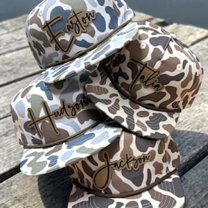 May include: A stack of camouflage print baseball caps with personalized names in a cursive font. The caps feature a rope detail and a curved brim. The names "Easton", "Hudson", "Talia", and "Jackson" are visible.