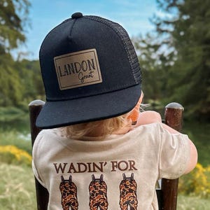 May include: A child wearing a black and brown camouflage print t-shirt with the text "Wadin' for Duck Season" and three cartoon figures wearing camouflage waders. The child is also wearing a black mesh trucker hat with a brown leather patch that says "Landon Grant".