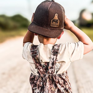 May include: A child wearing a brown trucker hat with the name "COLTON" on a leather patch, a cream-colored t-shirt, and camouflage overalls. The child is walking down a dirt road.