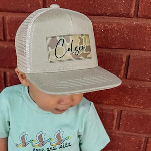 May include: A light blue t-shirt with a graphic of three ducks flying in formation and the text "free and wild". A child is wearing a beige and white trucker hat with a camouflage patch that says "Colsen" on it.
