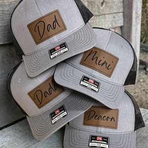 May include: A stack of gray trucker hats with black mesh backs. Each hat features a brown leather patch with a name: "Dad", "Mini", "Dada", and "Deacon". The hats have a Richardson 112 tag.