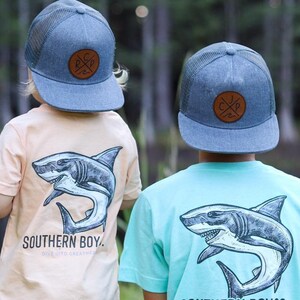 May include: Two children wearing blue and turquoise t-shirts with a shark graphic and the text "Southern Boyco. Dive into Greatness." They are both wearing blue mesh baseball caps with a brown leather patch.