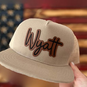 May include: A beige and black mesh trucker hat with a brown leather patch that says "Wyatt" in a cursive font.