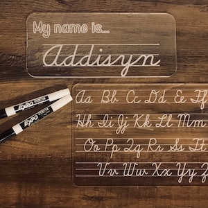 May include: Two clear acrylic dry-erase boards with white lines. The top board has the text "My name is..." and the name "Addisyn" written in cursive. The bottom board has the alphabet written in cursive in rows of five letters.
