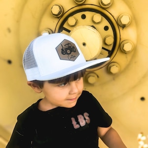 May include: A white mesh trucker hat with a brown leather patch featuring a tractor and the word "Cotton". A young person is wearing the hat and a black t-shirt with the word "LBS" on it.