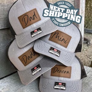 May include: A stack of gray trucker hats with black mesh backs and brown leatherette patches. Each hat has a different name: "Dad", "Mini", "Dada", and "Deacon". The hats have a Richardson 112 tag.