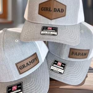 May include: Gray and white trucker hats with brown leatherette patches. The patches read "GIRL DAD", "GRAMPY", and "PAPAW". Each hat has a curved brim and an adjustable strap. The hats are from Richardson.