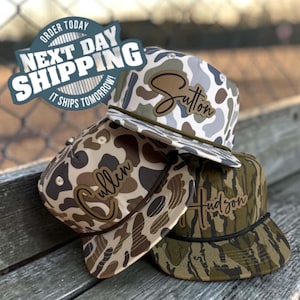 May include: Three baseball caps featuring camouflage patterns in shades of gray, brown, and green. Each cap has a brown leather-like patch on the front with a name engraved in cursive script: Sutton, Cullen, and Hudson. Each cap includes a rope trim.
