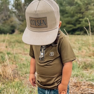 May include: A young person wearing a khaki mesh trucker hat with a brown leather patch that says "COLSEN" on the front. The person is also wearing a green short-sleeved polo shirt with a camouflage pattern on the collar.