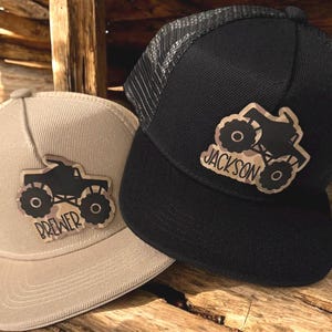 May include: Two trucker hats, one tan and one black, with a monster truck design. The tan hat has the name "BREWER" and the black hat has the name "JACKSON" on the patch. Both hats have a mesh back.