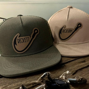 May include: Two trucker hats with a fishing hook design. One hat is olive green with a matching mesh back, and the other is tan. Both hats feature a brown patch with the name "BECKETT" in a curved font.