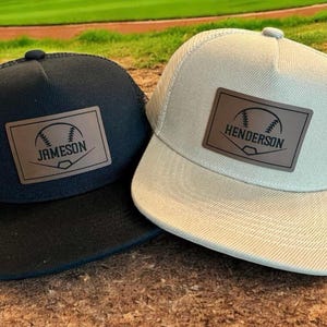 May include: Two baseball-themed trucker hats. One is black with a black mesh back and the other is tan with a white mesh back. Both have a brown leather patch with a baseball graphic and a name.