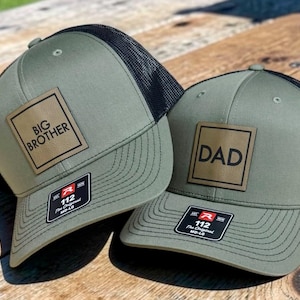 May include: Two olive green and black mesh trucker hats featuring a square leather-like patch on the front. One hat displays the text BIG BROTHER and the other displays the text DAD. Both hats have a black sticker on the brim with the text 112.