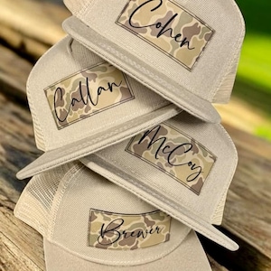 May include: A stack of four beige trucker hats with mesh backs. Each hat features a rectangular patch with a camouflage pattern and a name written in black script. The names visible on the patches are Cohen, Callan, McCoy, and Brewer.