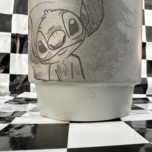 May include: A light gray ceramic planter featuring a detailed sketch of the character Stitch. The planter has a cylindrical shape with a slightly flared base. The artwork is in black and gray tones.