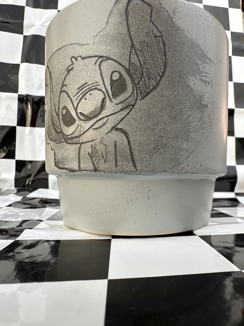 May include: A light gray ceramic planter featuring a detailed sketch of the character Stitch. The planter has a cylindrical shape with a slightly flared base. The artwork is in black and gray tones.
