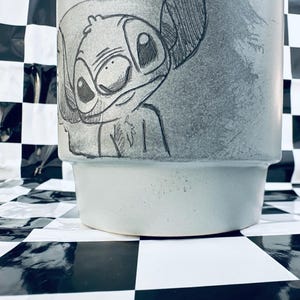 May include: A light blue ceramic planter featuring a hand-drawn sketch of the character Stitch. The sketch is in black and white, with detailed lines and shading. The planter has a wide, slightly flared shape.