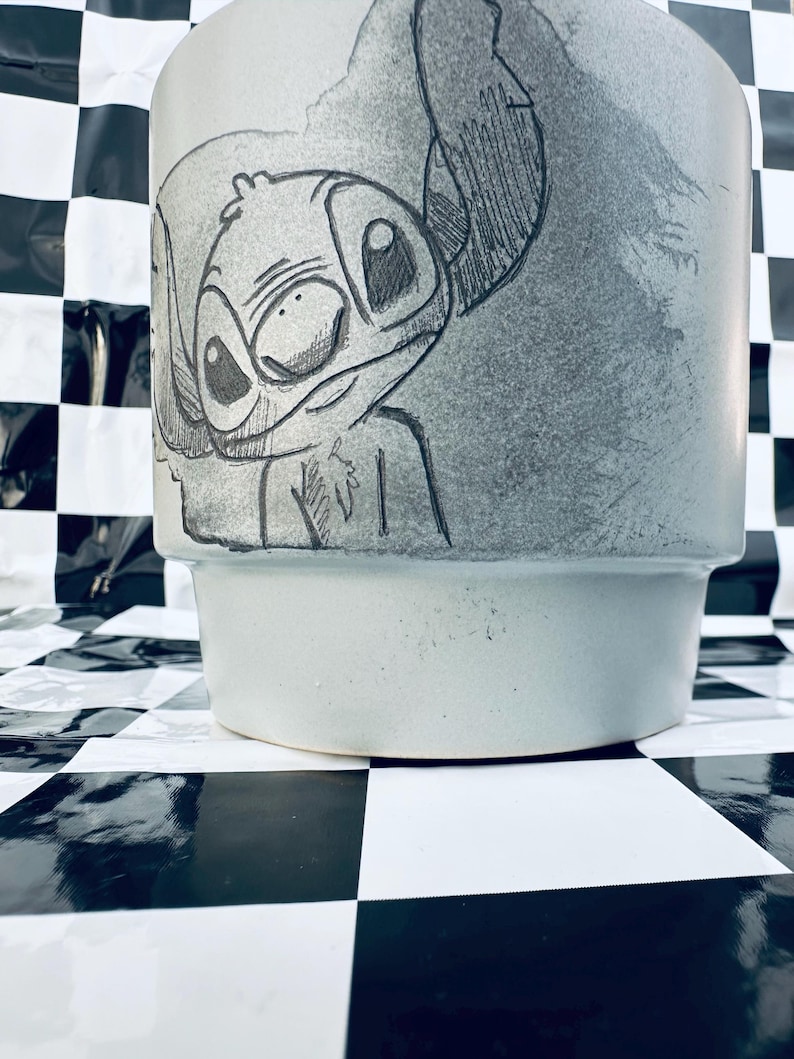 May include: A light blue ceramic planter featuring a hand-drawn sketch of the character Stitch. The sketch is in black and white, with detailed lines and shading. The planter has a wide, slightly flared shape.
