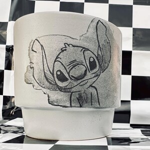 May include: A light gray ceramic planter featuring a hand-drawn illustration of Stitch from Lilo & Stitch. The artwork is in black and white, with Stitch's face and ears prominently displayed. The planter has a stepped design.