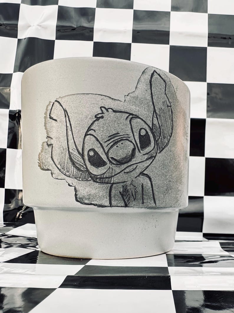 May include: A light gray ceramic planter featuring a hand-drawn illustration of Stitch from Lilo & Stitch. The artwork is in black and white, with Stitch's face and ears prominently displayed. The planter has a stepped design.