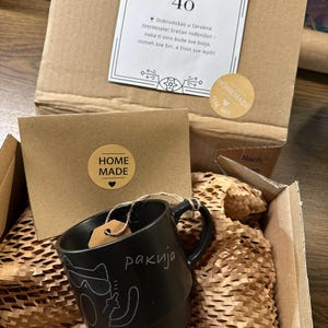 Puede incluir: Una taza de cer&aacute;mica negra con asa, con un dise&ntilde;o de mapache de dibujos animados y la palabra "rakija" en blanco. La taza est&aacute; dentro de una caja de cart&oacute;n con relleno de papel, una tarjeta peque&ntilde;a y una pegatina que dice "Homemade".