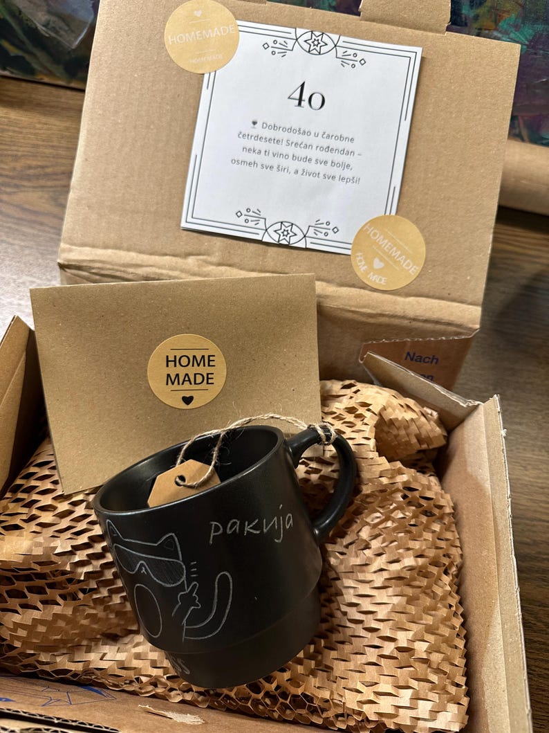 Puede incluir: Una taza de cer&aacute;mica negra con asa, con un dise&ntilde;o de mapache de dibujos animados y la palabra "rakija" en blanco. La taza est&aacute; dentro de una caja de cart&oacute;n con relleno de papel, una tarjeta peque&ntilde;a y una pegatina que dice "Homemade".