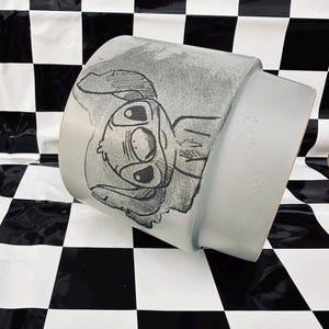 May include: A light gray ceramic mug featuring a black and white illustration of a cartoon character. The mug has a cylindrical shape with a slightly flared top. The character's face is prominently displayed on the side of the mug.