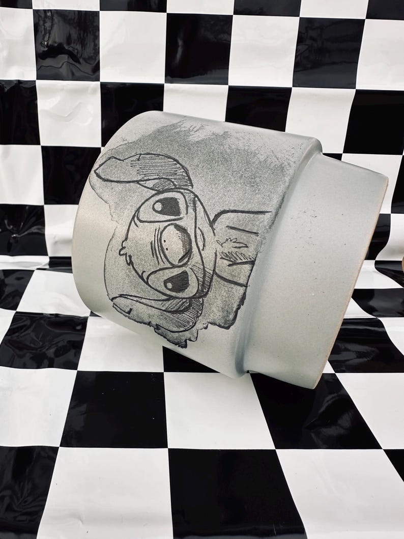 May include: A light gray ceramic mug featuring a black and white illustration of a cartoon character. The mug has a cylindrical shape with a slightly flared top. The character's face is prominently displayed on the side of the mug.