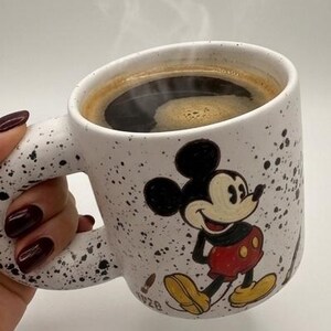 Puede incluir: Taza de caf&eacute; de cer&aacute;mica blanca con un dise&ntilde;o de Mickey Mouse en negro y rojo. La taza tiene un patr&oacute;n moteado y est&aacute; llena de caf&eacute; caliente, con vapor saliendo. El asa tiene una forma curva &uacute;nica.