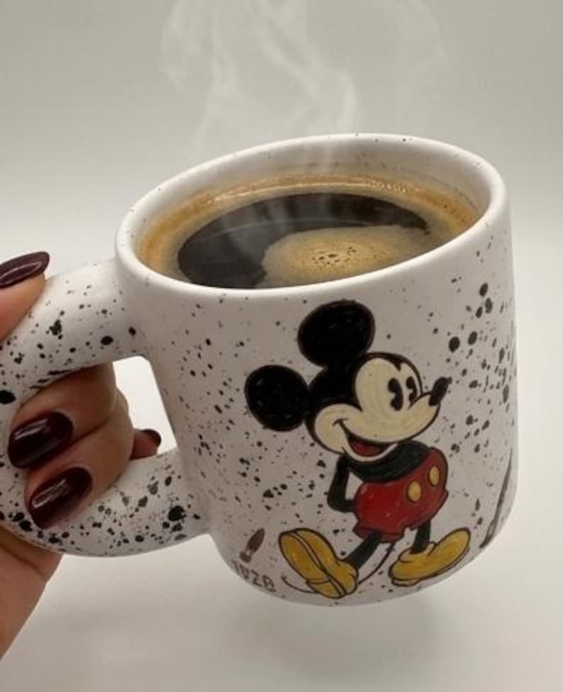 Puede incluir: Taza de caf&eacute; de cer&aacute;mica blanca con un dise&ntilde;o de Mickey Mouse en negro y rojo. La taza tiene un patr&oacute;n moteado y est&aacute; llena de caf&eacute; caliente, con vapor saliendo. El asa tiene una forma curva &uacute;nica.