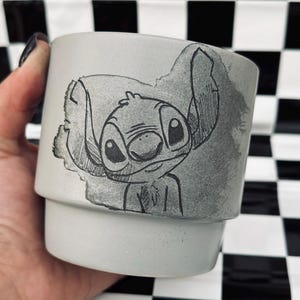 May include: A light gray ceramic mug featuring a hand-drawn illustration of Stitch from Lilo & Stitch. The artwork is in black and gray tones. The mug has a cylindrical shape with a slightly flared base, perfect for holding beverages.