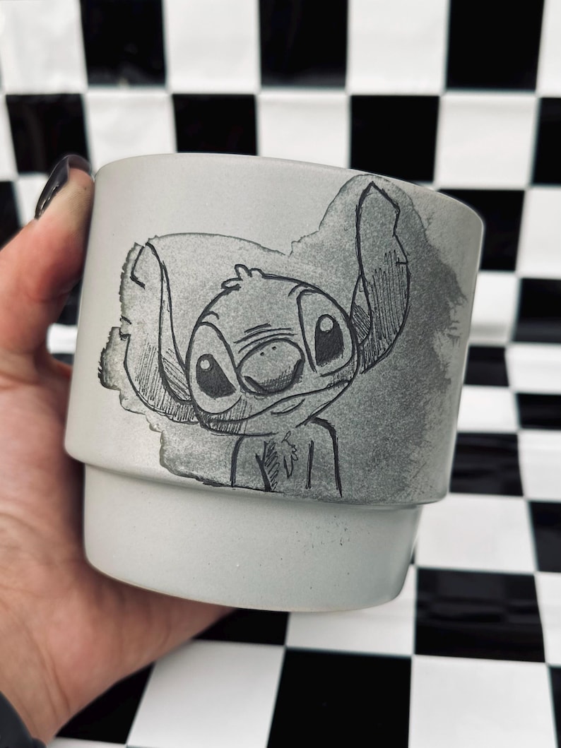 May include: A light gray ceramic mug featuring a hand-drawn illustration of Stitch from Lilo & Stitch. The artwork is in black and gray tones. The mug has a cylindrical shape with a slightly flared base, perfect for holding beverages.
