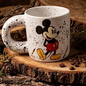 Puede incluir: Taza de cer&aacute;mica blanca con dise&ntilde;o moteado y asa grande y redondeada. La taza presenta una colorida ilustraci&oacute;n de Mickey Mouse. El a&ntilde;o "1928" est&aacute; impreso en la taza. Granos de caf&eacute; y una pi&ntilde;a se encuentran sobre una superficie de madera.