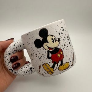 Puede incluir: Taza de cer&aacute;mica blanca con dise&ntilde;o moteado negro y un asa grande y curvada. La taza presenta una imagen pintada a mano de Mickey Mouse con su atuendo cl&aacute;sico, con el a&ntilde;o "1928" impreso debajo. Tambi&eacute;n hay pinceles pintados en la taza.