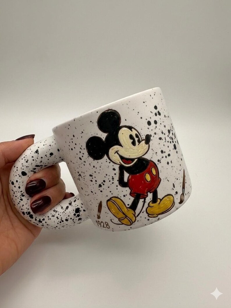 Puede incluir: Taza de cer&aacute;mica blanca con dise&ntilde;o moteado negro y un asa grande y curvada. La taza presenta una imagen pintada a mano de Mickey Mouse con su atuendo cl&aacute;sico, con el a&ntilde;o "1928" impreso debajo. Tambi&eacute;n hay pinceles pintados en la taza.