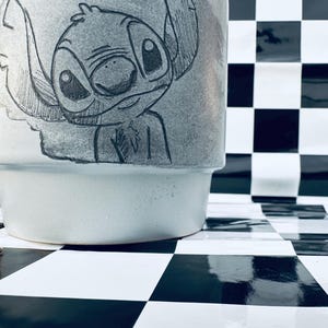 May include: A light gray ceramic mug featuring a hand-drawn illustration of the character Stitch from the Disney movie Lilo & Stitch. The mug has a wide base and a slightly flared top. The background is a black and white checkered pattern.