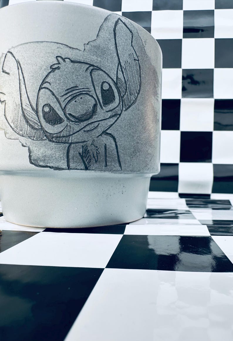 May include: A light gray ceramic mug featuring a hand-drawn illustration of the character Stitch from the Disney movie Lilo & Stitch. The mug has a wide base and a slightly flared top. The background is a black and white checkered pattern.