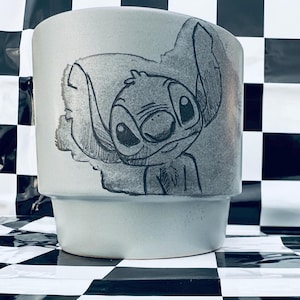 May include: A light gray ceramic planter featuring a hand-drawn illustration of the character Stitch. The artwork is in black and gray, with a detailed depiction of Stitch's face and ears. The planter has a cylindrical shape with a slightly flared base.