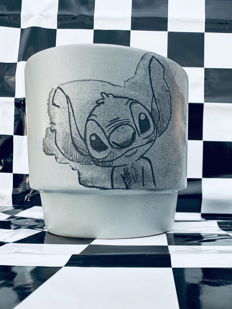 May include: A light gray ceramic planter featuring a hand-drawn illustration of the character Stitch. The artwork is in black and gray, with a detailed depiction of Stitch's face and ears. The planter has a cylindrical shape with a slightly flared base.