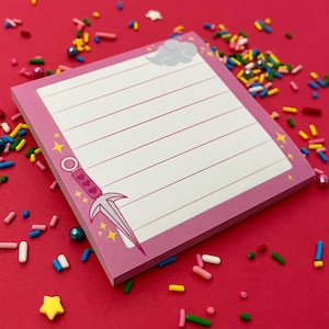 May include: Pink square notepad with lined pages, a decorative border, and a cartoon dagger illustration. The notepad is surrounded by colourful sprinkles on a red background. The notepad has a cloud illustration in the top corner.