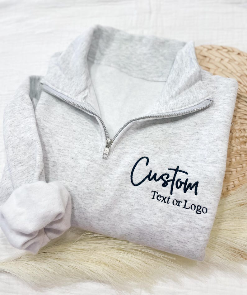 May include: A gray quarter-zip sweatshirt with a black embroidered design that says "Custom Text or Logo".
